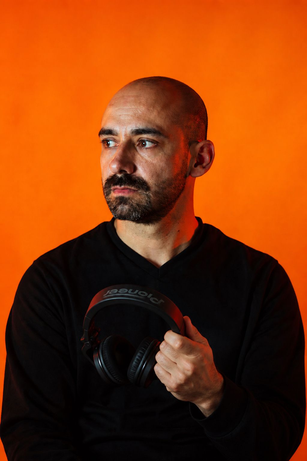 XPLCT! holding Pioneer DJ headphones against an orange studio backdrop